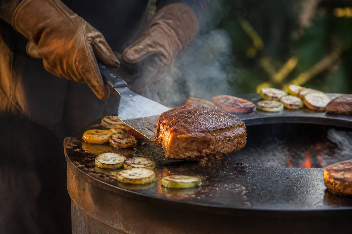 Mains gantées retournant une pièce de viande à la spatule sur la couronne d’acier chaude d’un brasero